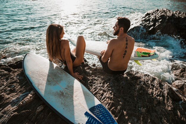 Mujer joven y hombre con tablas de surf sentado en la roca cerca del mar