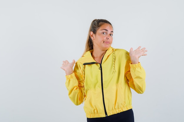 Mujer joven en impermeable amarillo mostrando gesto de impotencia mientras se encoge de hombros