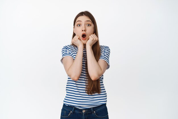 Mujer joven impresionada mirando con asombro con la mandíbula caída, mirando emocionado, tomarse de las manos cerca de la cara con emoción de sorpresa en blanco.