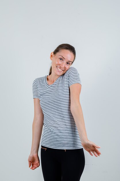 Mujer joven inclinando la cabeza sobre el hombro en camiseta, pantalón y mirando optimista, vista frontal.