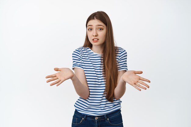 Mujer joven incómoda que muestra las manos vacías y se encoge de hombros, no tiene nada, despistada contra el fondo blanco.