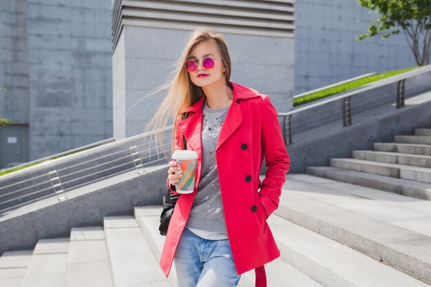 Mujer joven inconformista en abrigo rosa, jeans en la calle con café escuchando música en auriculares, con gafas de sol