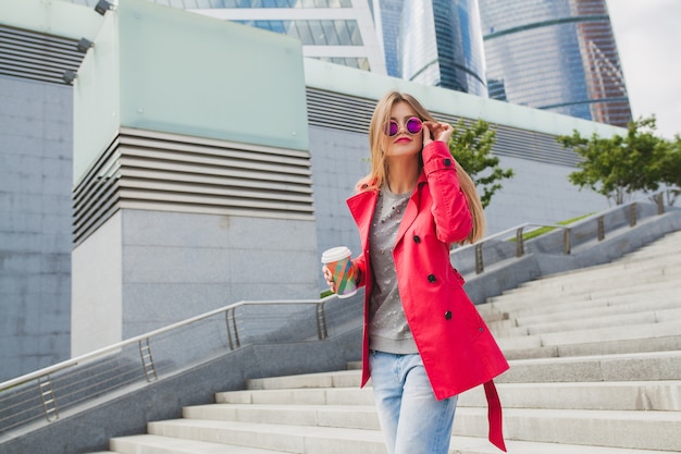 Mujer joven inconformista en abrigo rosa, jeans en la calle con café escuchando música en auriculares, con gafas de sol