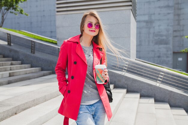 Mujer joven inconformista en abrigo rosa, jeans en la calle con café escuchando música en auriculares