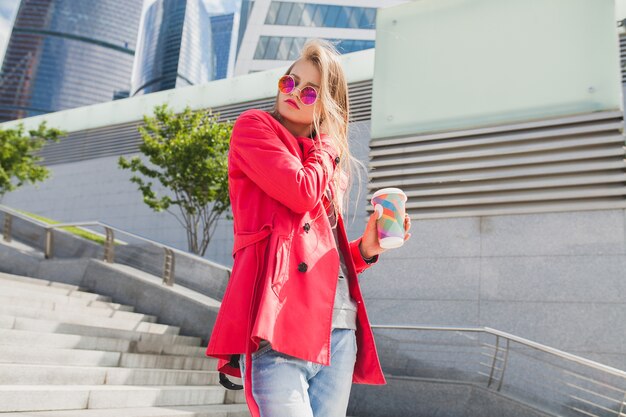 Mujer joven inconformista en abrigo rosa, jeans en la calle con café escuchando música en auriculares