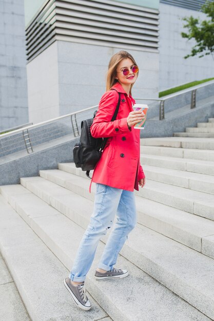 Mujer joven inconformista en abrigo rosa, jeans en la calle con mochila y café escuchando música en auriculares, con gafas de sol