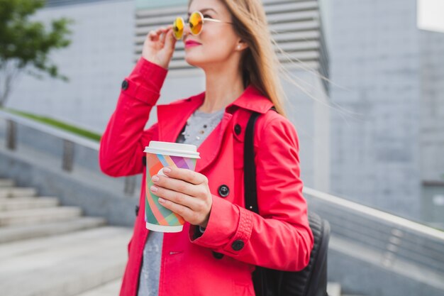 Mujer joven inconformista en abrigo rosa, jeans en la calle con mochila y café escuchando música en auriculares