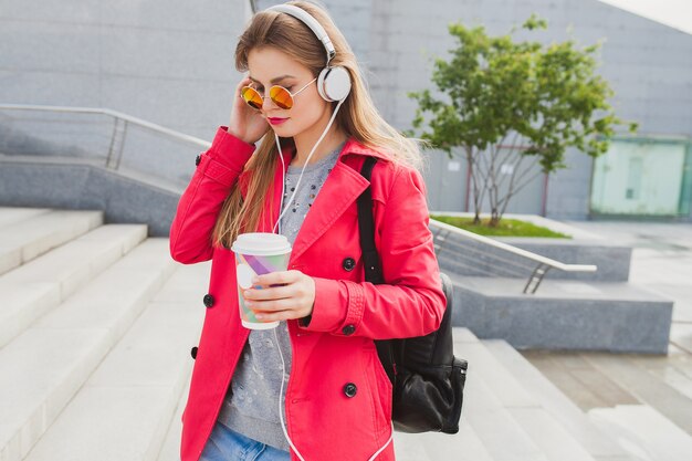 Mujer joven inconformista en abrigo rosa, jeans en la calle con mochila y café escuchando música en auriculares