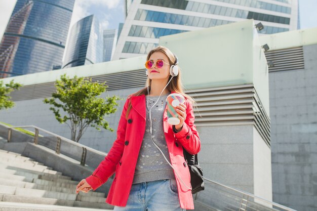 Mujer joven inconformista en abrigo rosa, jeans en la calle con mochila y café escuchando música en auriculares