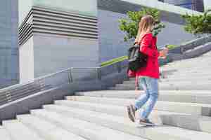 Foto gratuita mujer joven inconformista en abrigo rosa, jeans caminando por las escaleras en la calle con mochila y café escuchando música en auriculares, con gafas de sol