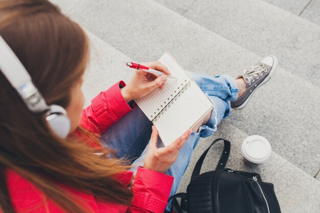 Mujer joven inconformista en abrigo rosa, jeans sentado en la calle con mochila y café escuchando música en auriculares