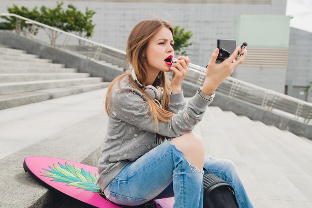 Mujer joven inconformista en la calle haciendo maquillaje lápiz labial rojo sentado con tabla de equilibrio vistiendo suéter y jeans