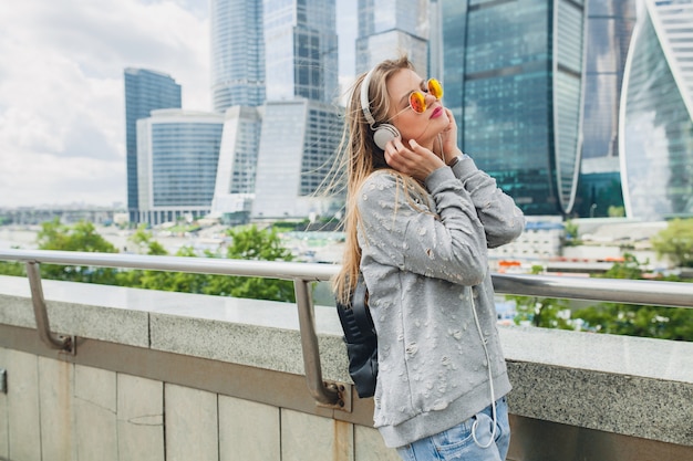 Mujer joven inconformista divirtiéndose en la calle escuchando música en auriculares, con gafas de sol rosa