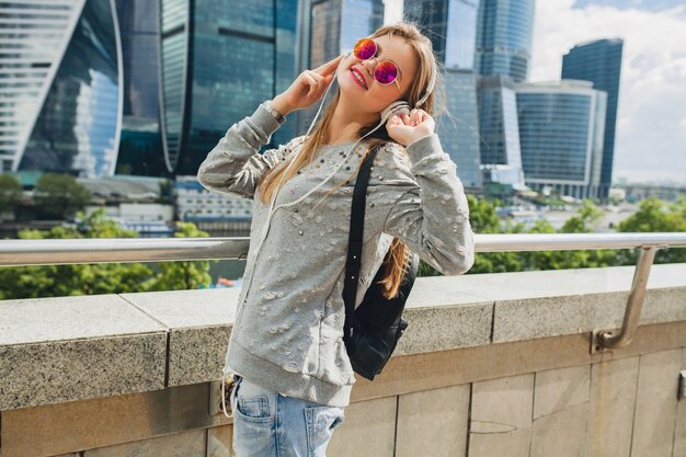 Mujer joven inconformista divirtiéndose en la calle escuchando música en auriculares, con gafas de sol rosa