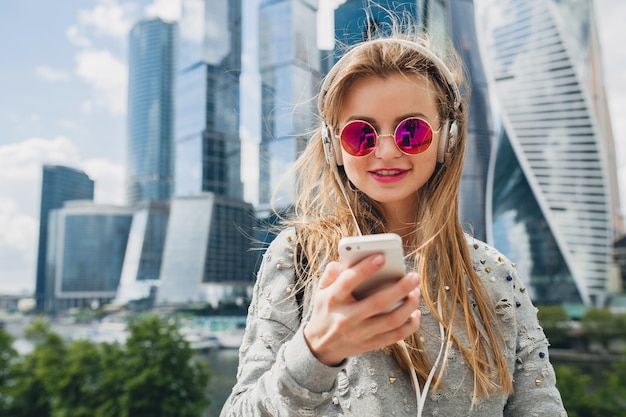 Mujer joven inconformista divirtiéndose en la calle escuchando música en auriculares, con gafas de sol rosas, estilo urbano primavera verano, con smartphone