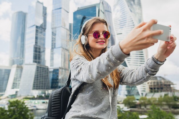 Mujer joven inconformista divirtiéndose en la calle escuchando música en auriculares, con gafas de sol rosas, estilo urbano primavera verano, tomando selfie pisture en smartphone