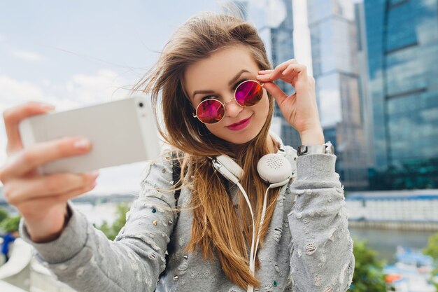 Mujer joven inconformista divirtiéndose en la calle, con gafas de sol rosas, estilo urbano primavera verano, tomando selfie pisture en smartphone