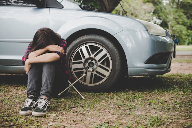 La mujer joven del inconformista que espera para la ayuda del borde de la carretera después de su coche se rompe en el lado de la carretera que se sienta contra su coche.