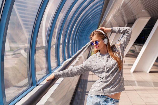 Mujer joven inconformista en ropa casual divirtiéndose escuchando música en auriculares, vistiendo jeans, suéter y gafas de sol, estilo urbano