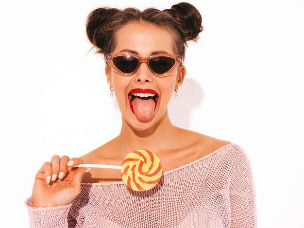 Mujer joven inconformista sexy hermosa con labios rojos en gafas de sol. Comer, morder paleta de caramelo. Muestra lengua