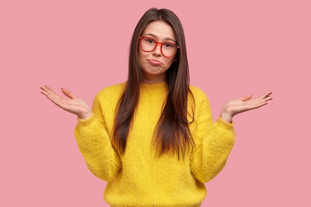 Mujer joven indecisa vacilante frunce el labio inferior, extiende las palmas de las manos con expresión despistada, vestida con un jersey amarillo cálido informal, sin darse cuenta