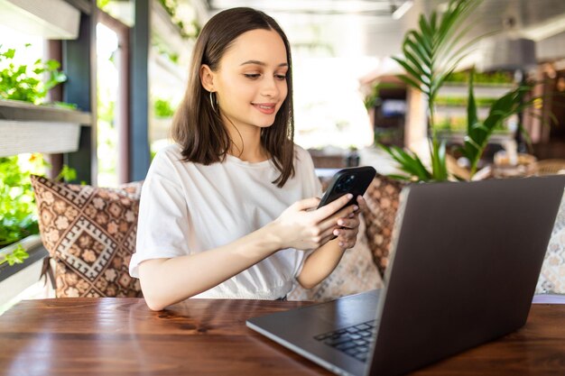 Mujer joven independiente sentada en la cafetería con un portátil y tomando café mientras usa su teléfono