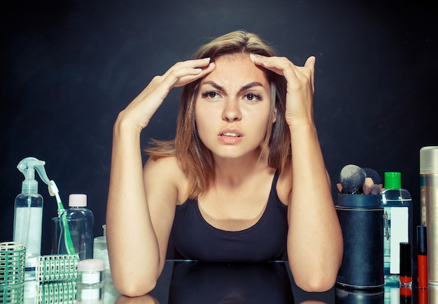 Mujer joven infeliz insatisfecha mirando a sí misma en el espejo sobre fondo negro de estudio. concepto de piel y acné roblem. Conceptos de mañana, maquillaje y emociones humanas.