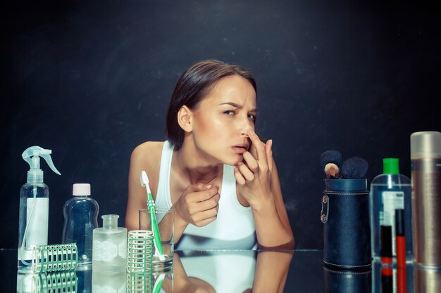 Mujer joven infeliz insatisfecha mirando a sí misma en el espejo sobre fondo negro de estudio. piel de roblem y concepto de acné modelo caucásico en el estudio