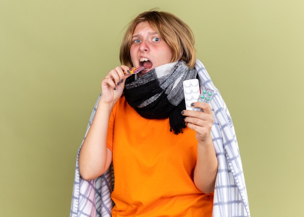 Mujer joven insalubre en camiseta naranja con bufanda caliente alrededor del cuello malestar que sufre de gripe sosteniendo diferentes píldoras que parecen confundidas
