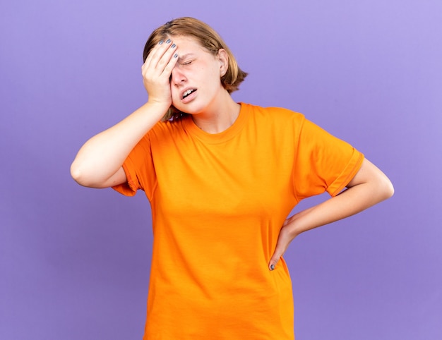 Mujer joven insalubre en camiseta naranja que se siente mal tocando su frente mientras se siente mareado con gripe de pie sobre la pared púrpura