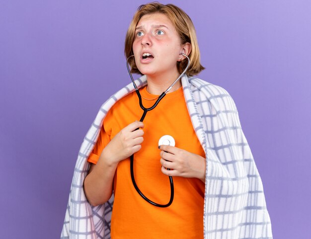 Mujer joven insalubre envuelta en una manta caliente sintiéndose enferma escuchando los latidos de su corazón con estetoscopio mirando preocupado