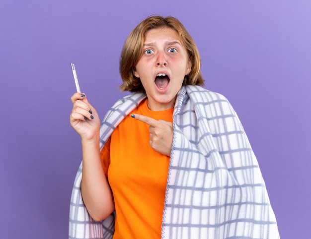Mujer joven insalubre envuelta en una manta caliente sintiéndose enferma que sufre de gripe que tiene fiebre midiendo su temperatura con termómetro mirando preocupado