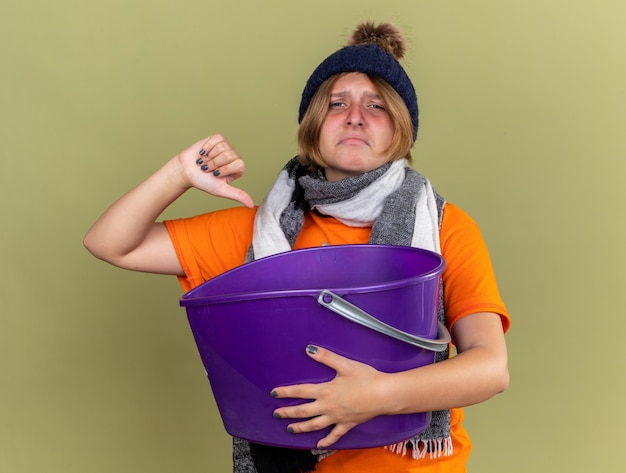 Mujer joven insalubre con sombrero con bufanda alrededor de su cuello sintiéndose enferma sufriendo de náuseas sosteniendo el lavabo mostrando los pulgares hacia abajo de pie sobre la pared verde