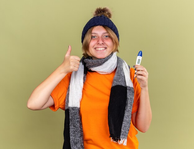 Mujer joven insalubre con sombrero con bufanda alrededor de su cuello sintiéndose mejor sosteniendo el termómetro digital sonriendo mostrando los pulgares para arriba de pie sobre la pared verde