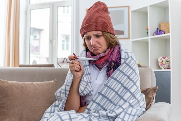 Mujer joven insalubre con sombrero con bufanda caliente alrededor del cuello con termómetro comprobando su temperatura sintiéndose mal que sufre de resfriado y gripe con aspecto preocupado sentado en el sofá en la sala de estar luminosa