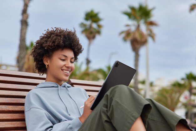 Mujer joven de inspiración positiva con poses de cabello rizado en un banco de madera usa tableta digital y lápiz óptico hace bocetos tiene expresión feliz pasa tiempo libre en el aire fresco Concepto de estilo de vida de la gente