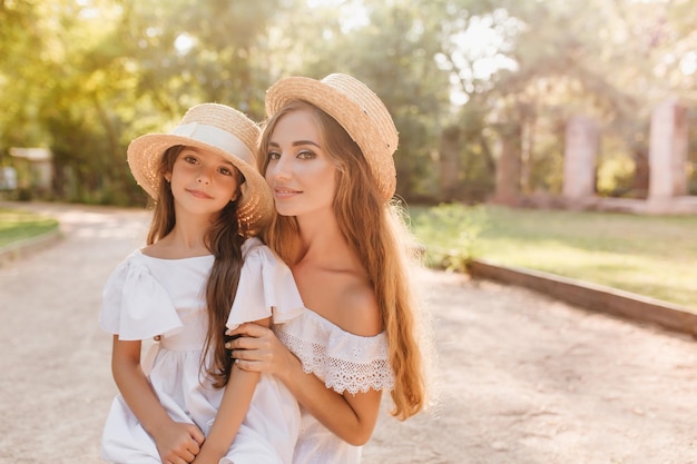 Mujer joven inspirada con bronceado hermoso que presenta con placer junto a la pequeña hija en traje elegante. Madre atractiva sonriendo suavemente abrazando al niño en el callejón soleado en la mañana.