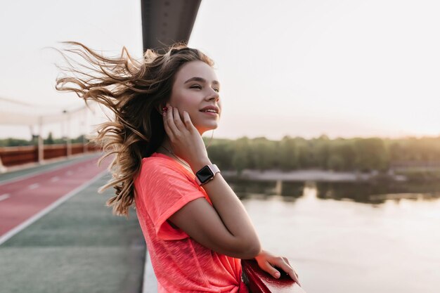 Mujer joven inspirada con cabello ondulado posando cerca del lago Chica relajada con reloj inteligente de pie sobre el fondo de la naturaleza en la mañana