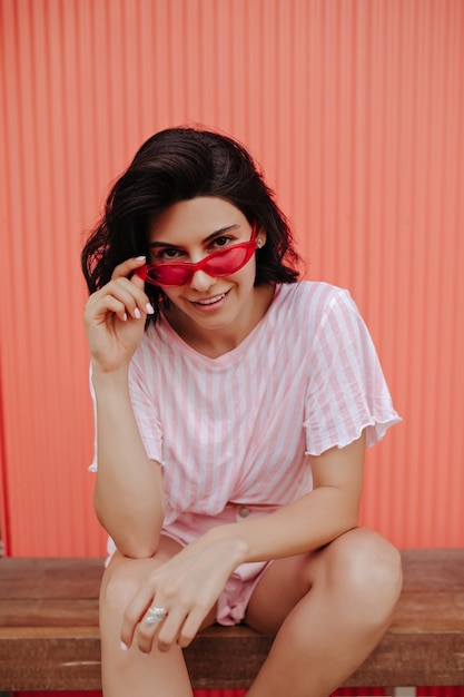 Foto gratuita mujer joven interesada mirando a través de gafas de sol rosas. disparo al aire libre de una mujer bonita morena sentada en un banco de madera.