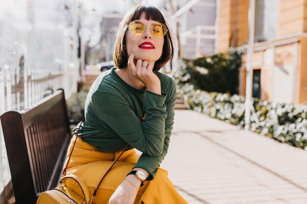 Mujer joven interesada en pantalones amarillos sentado en la calle. Foto al aire libre de una hermosa chica morena posando en un banco.