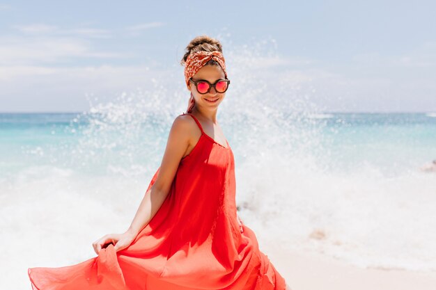 Mujer joven interesada en traje de verano con estilo divirtiéndose cerca del océano. Retrato al aire libre de adorable niña caucásica bailando en la naturaleza.