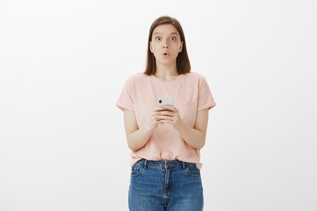 Mujer joven intrigada doblando los labios asombrados después de leer el mensaje en el teléfono inteligente, usando la aplicación de teléfono móvil