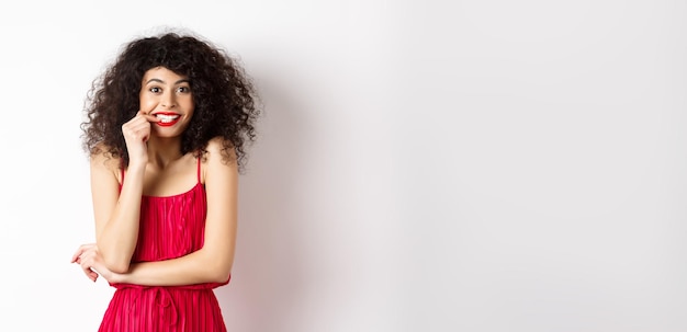 Foto gratuita mujer joven intrigada con el pelo rizado con un elegante vestido rojo y pintalabios mordiéndose las uñas y