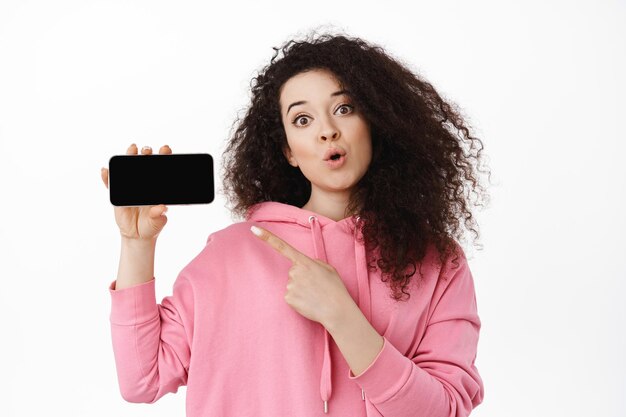 Mujer joven intrigada señalando la pantalla en blanco del teléfono móvil, mostrando una interesante promoción en línea, aplicación en el teléfono inteligente, de pie en una sudadera con capucha contra un fondo blanco.