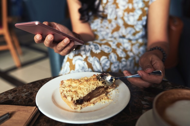 Mujer joven irreconocible sentada en la cafetería, usando un teléfono inteligente y comiendo pastel de crumble