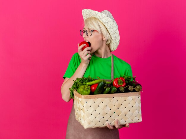 Mujer joven jardinero con pelo corto en delantal y sombrero con caja llena de verduras