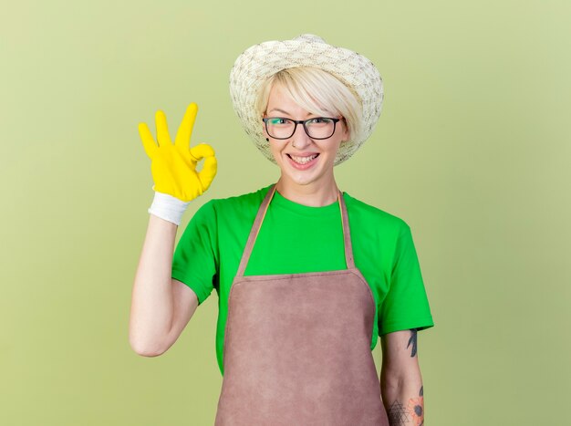 Mujer joven jardinero con pelo corto en delantal y sombrero con guantes de goma mirando a la cámara sonriendo con cara feliz mostrando signo ok de pie sobre fondo claro