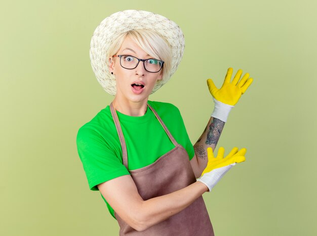 Mujer joven jardinero con pelo corto en delantal y sombrero con guantes de goma presentando algo con los brazos mirando sorprendido y confundido de pie sobre fondo claro
