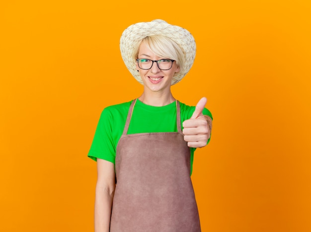 Mujer joven jardinero con pelo corto en delantal y sombrero mirando a la cámara sonriendo con cara feliz mostrando los pulgares para arriba sobre fondo naranja