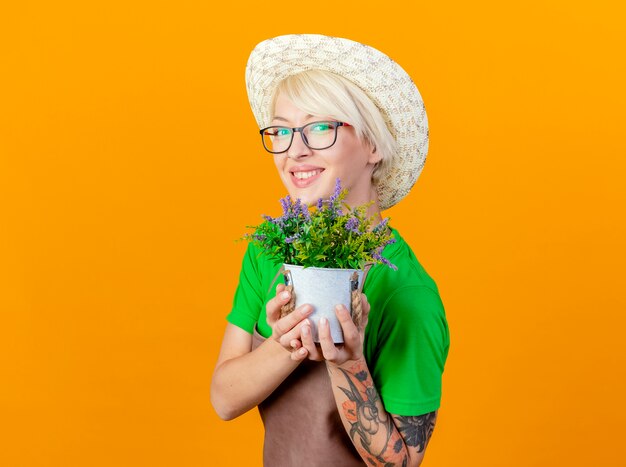 Mujer joven jardinero con pelo corto en delantal y sombrero mostrando planta en maceta mirando a cámara sonriendo con cara feliz de pie sobre fondo naranja